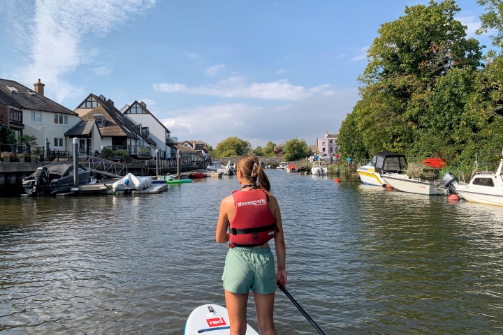 Person paddleboarding on a calm river with houses and boats on either side.